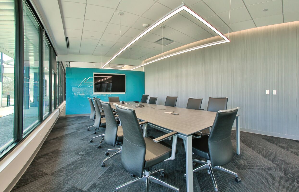 Large conference room with U‑shaped tables, ergonomic chairs, multiple wall‑mounted displays, and linear ceiling lighting adjacent to a glass‑lined corridor.