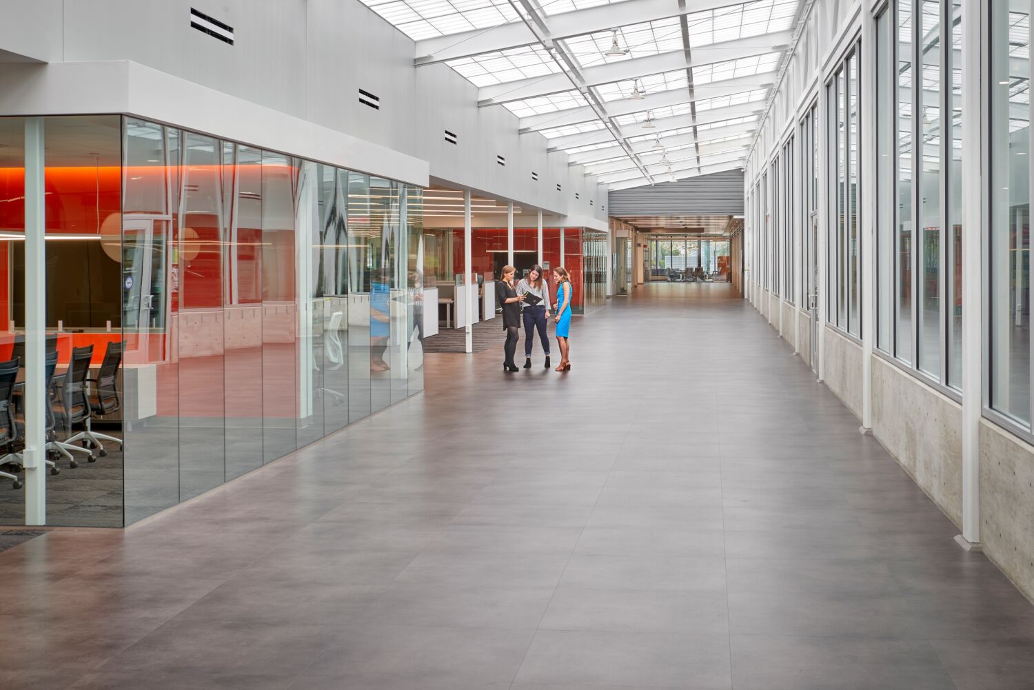 Wide interior corridor with a glass‑walled meeting room featuring orange accent lighting, skylight ceiling panels, and a small group of people standing and talking.