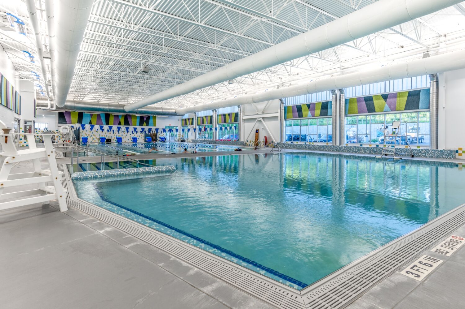 Indoor swimming pool area with lap lanes, colorful upper wall panels, large windows, and an exposed structural ceiling.