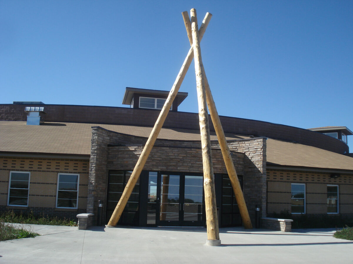 Exterior entrance of a lodge‑style building featuring a tall wood‑pole structural element, stone façade, and large glass doors under a clear sky.