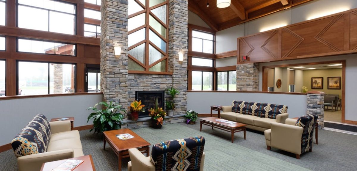 Lodge‑style community living room with stone fireplace, wood‑beam ceiling, large windows, and seating arranged around central tables.