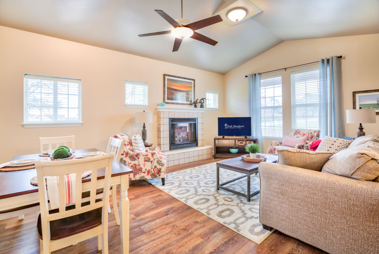Living room at Park Meadows Senior Living with seating area, fireplace, dining table, ceiling fan, and hardwood flooring.