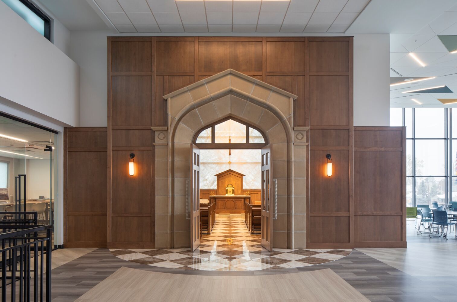 Chapel entrance with a stone gothic-style archway, wood-paneled wall surround, double doors, and a view into the chapel featuring a central altar and patterned flooring.