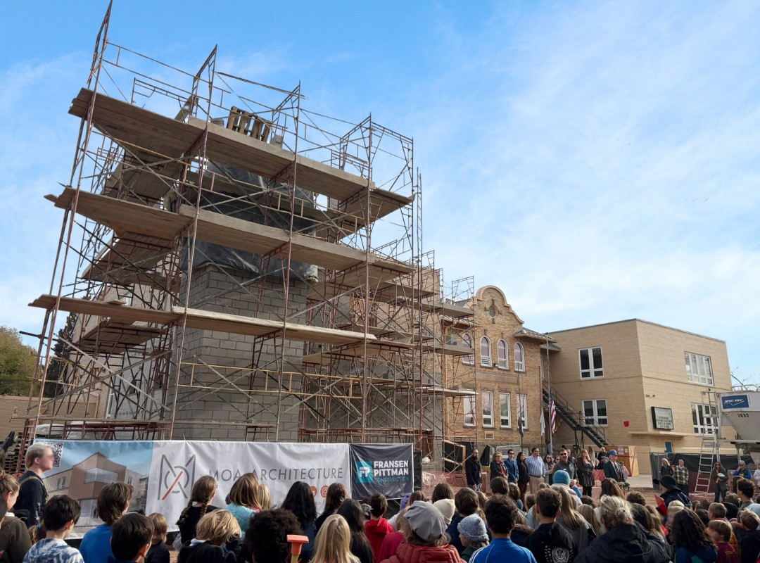 Construction site of a school building with multi‑story scaffolding, masonry walls in progress, historic brick structures in the background, and banners displaying project partners along the fenced perimeter.