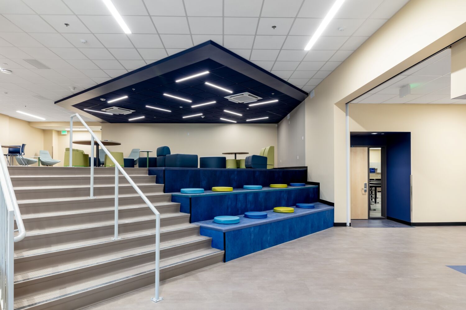 Tiered collaboration area with built‑in stadium‑style seating and round floor cushions, adjacent stairs with metal handrails, overhead ceiling panels with integrated linear lighting, and nearby lounge tables and chairs.