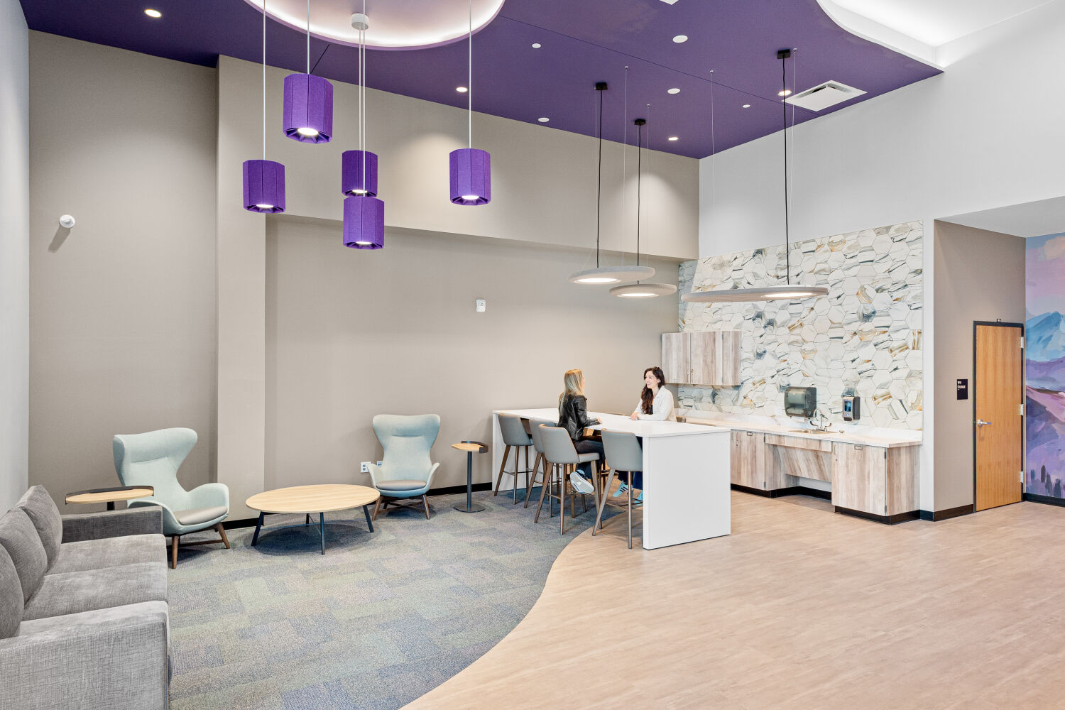 Community room with lounge seating, a kitchenette island with stools, pendant lighting, patterned wall tile, and a mix of carpet and wood‑look flooring.