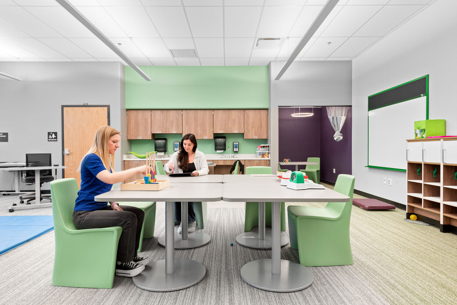 Behavioral classroom with grouped worktables, flexible seating, perimeter cabinetry with sinks, adjacent therapy mat area, and a partially enclosed calming nook.