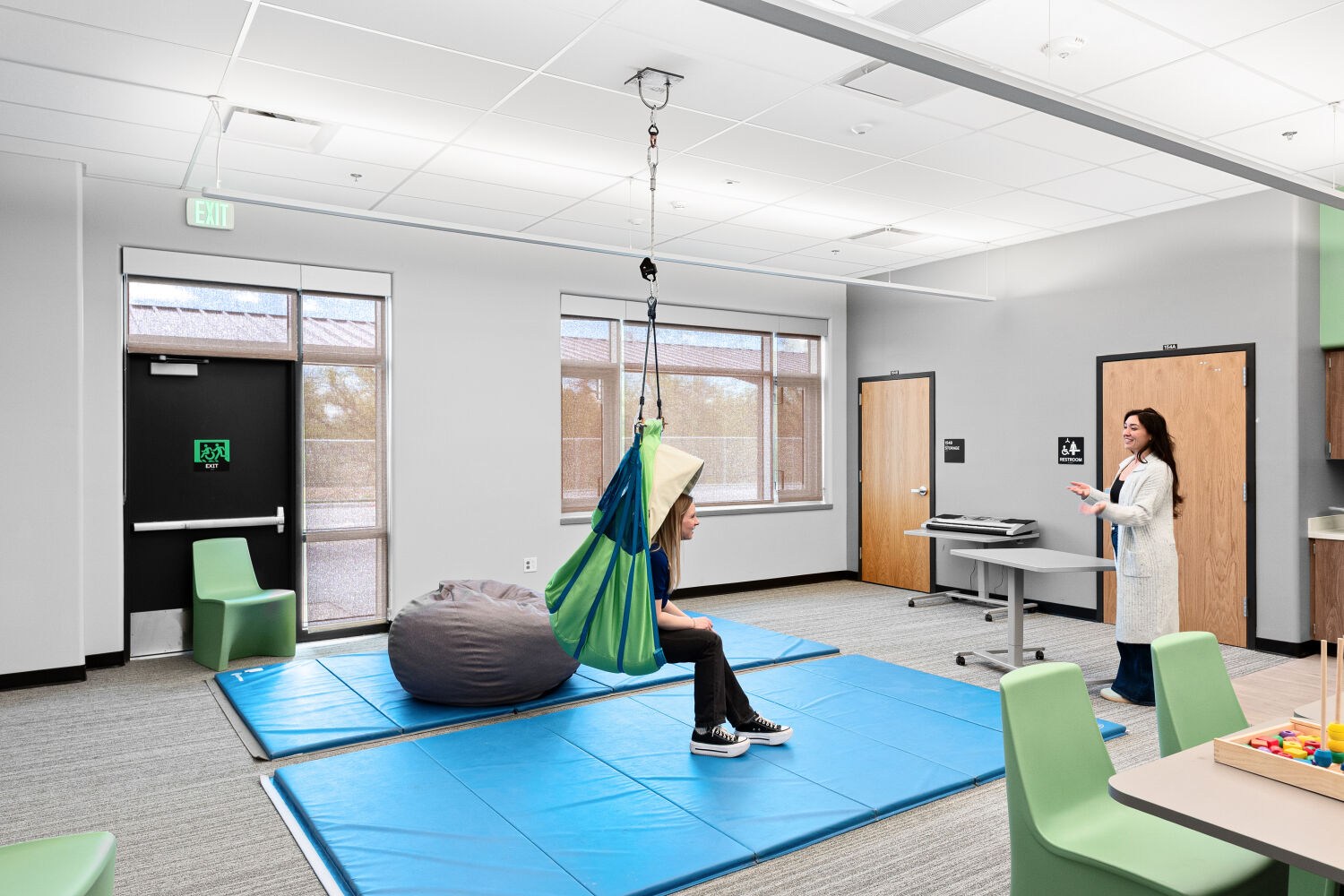 Behavioral needs classroom with a ceiling‑mounted lift system, floor mats, soft seating, mobile work table, perimeter windows, and resilient flooring.