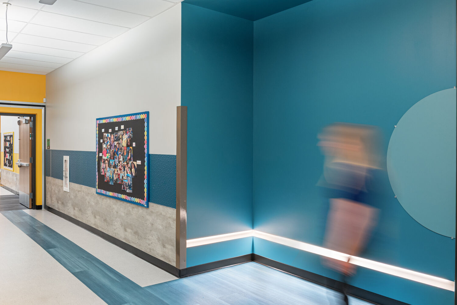 Learning corridor with blue accent walls, resilient flooring with color inlays, integrated wall lighting, classroom entry doors, and student display boards.