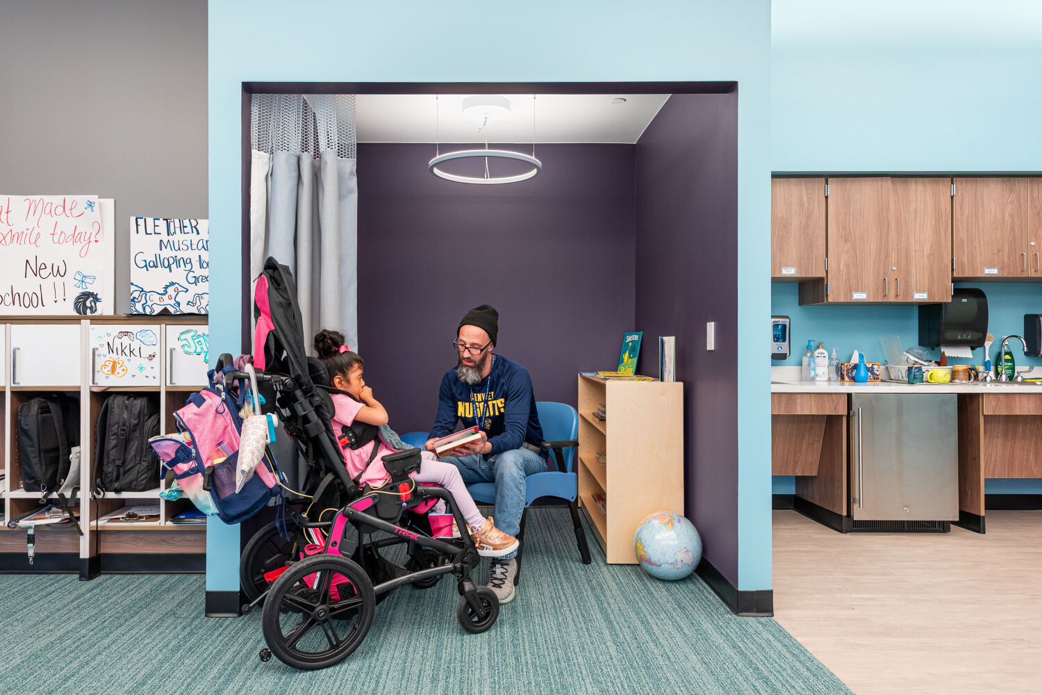 Small reading nook with an alcove seating area, low book shelving, soft flooring, circular pendant light, and adjacent classroom storage cabinets.