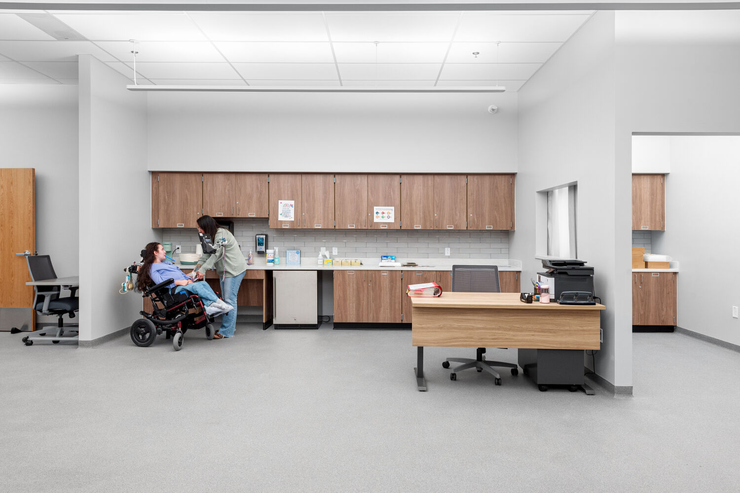 School clinic with perimeter cabinetry and sink, medical supply storage, open care area, staff work desk, and adjacent exam and support rooms.