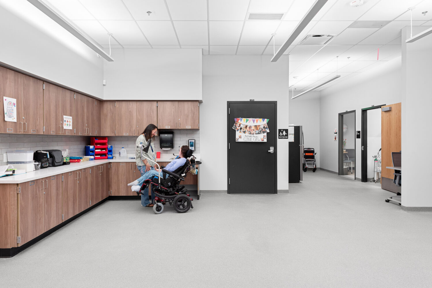 School clinic with perimeter cabinetry and sink, medical storage, open floor space for care activities, and adjacent support rooms.