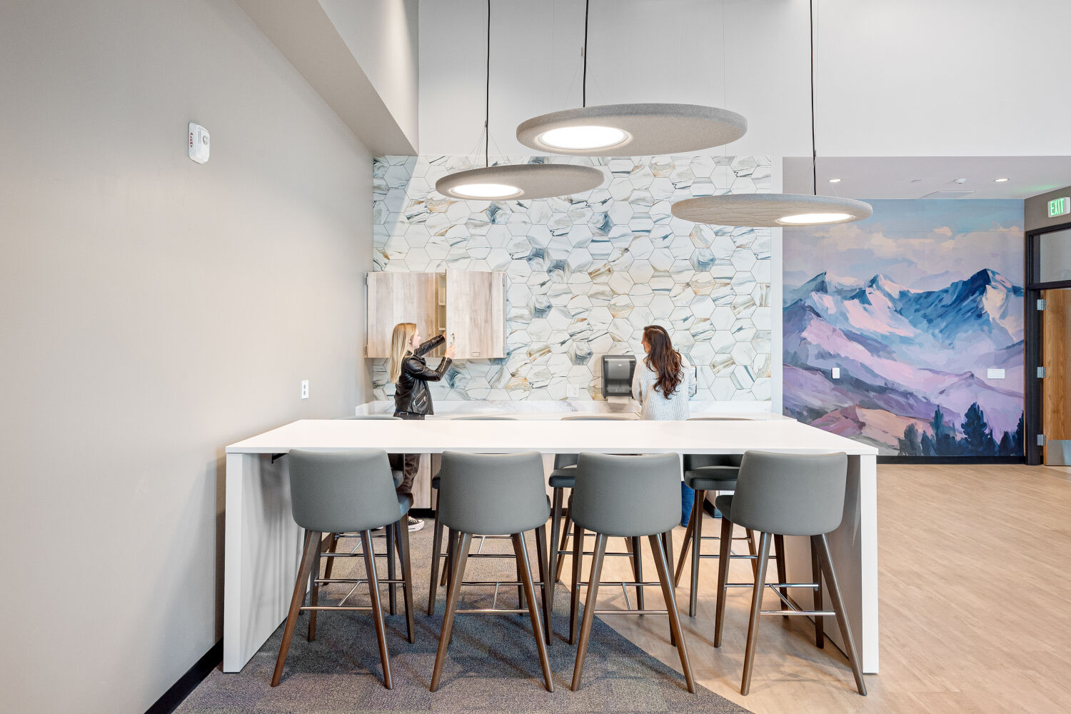 Community room high-top seating with a long island and stools, pendant lighting, patterned tile backsplash, wood cabinetry, and adjacent lounge seating.