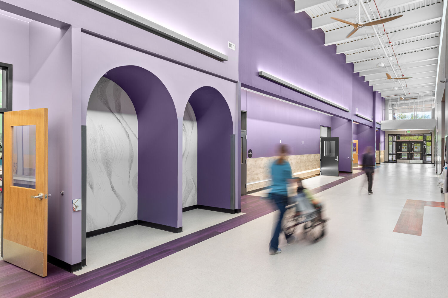 Main school corridor with purple accent walls, arched wall niches, built‑in bench seating, resilient flooring, and a high ceiling with exposed structure and linear lighting.