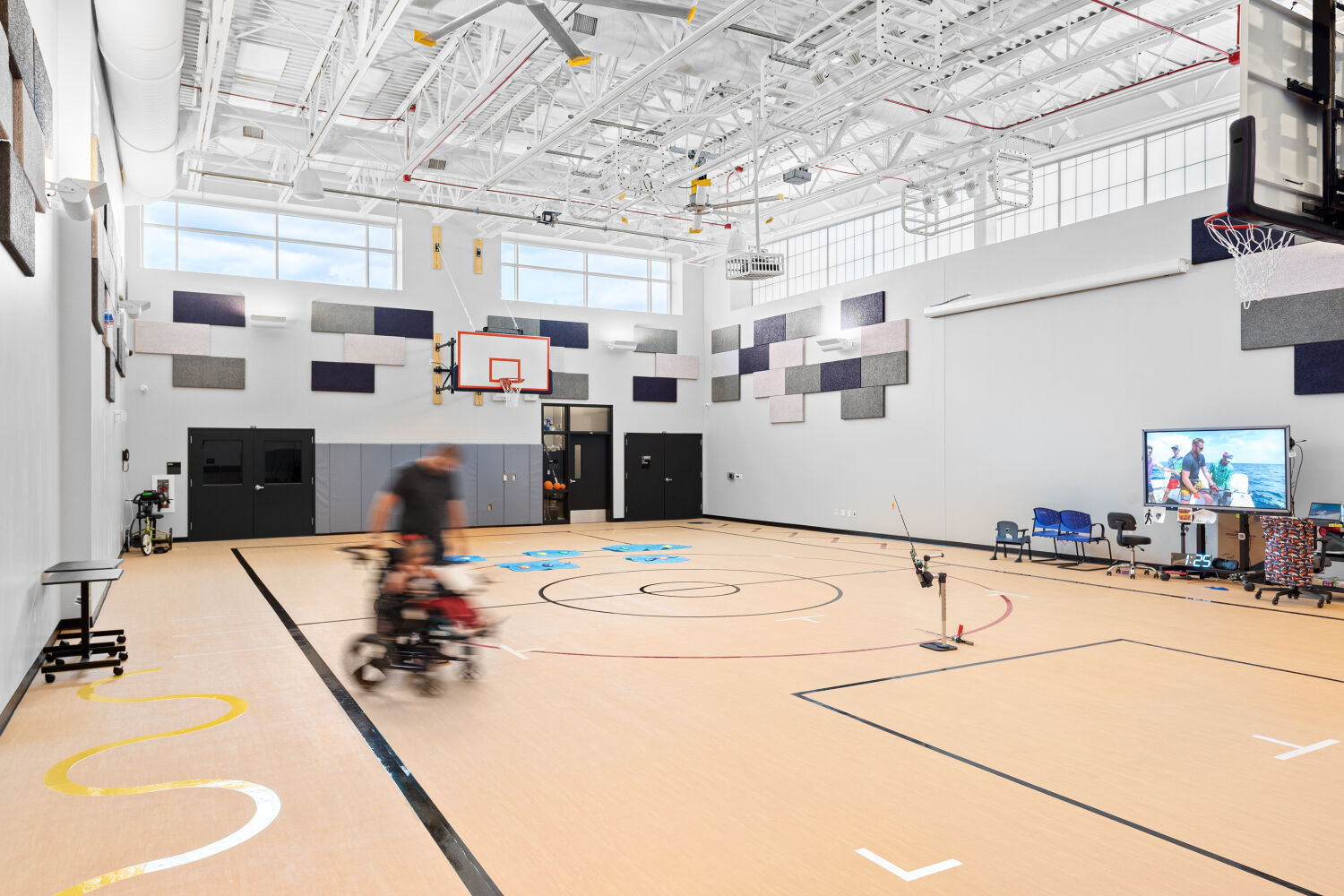 School gymnasium with a multi‑court floor layout, wall‑mounted basketball hoops, acoustic wall panels, high exposed roof structure, and large perimeter windows.