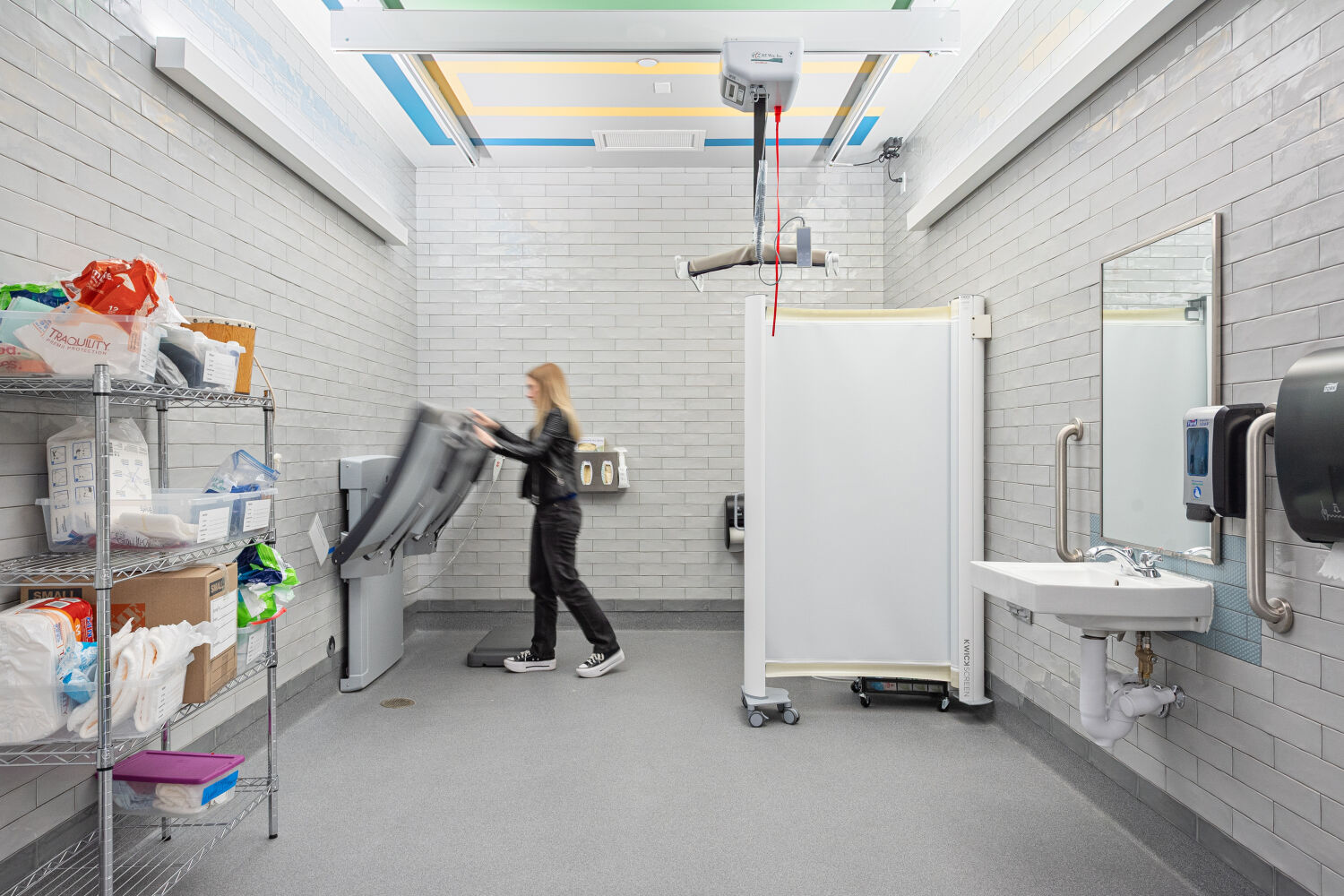 Hygiene room with a ceiling‑mounted lift system, height‑adjustable care table, privacy screen, wall‑mounted sink, open supply shelving, and resilient flooring.