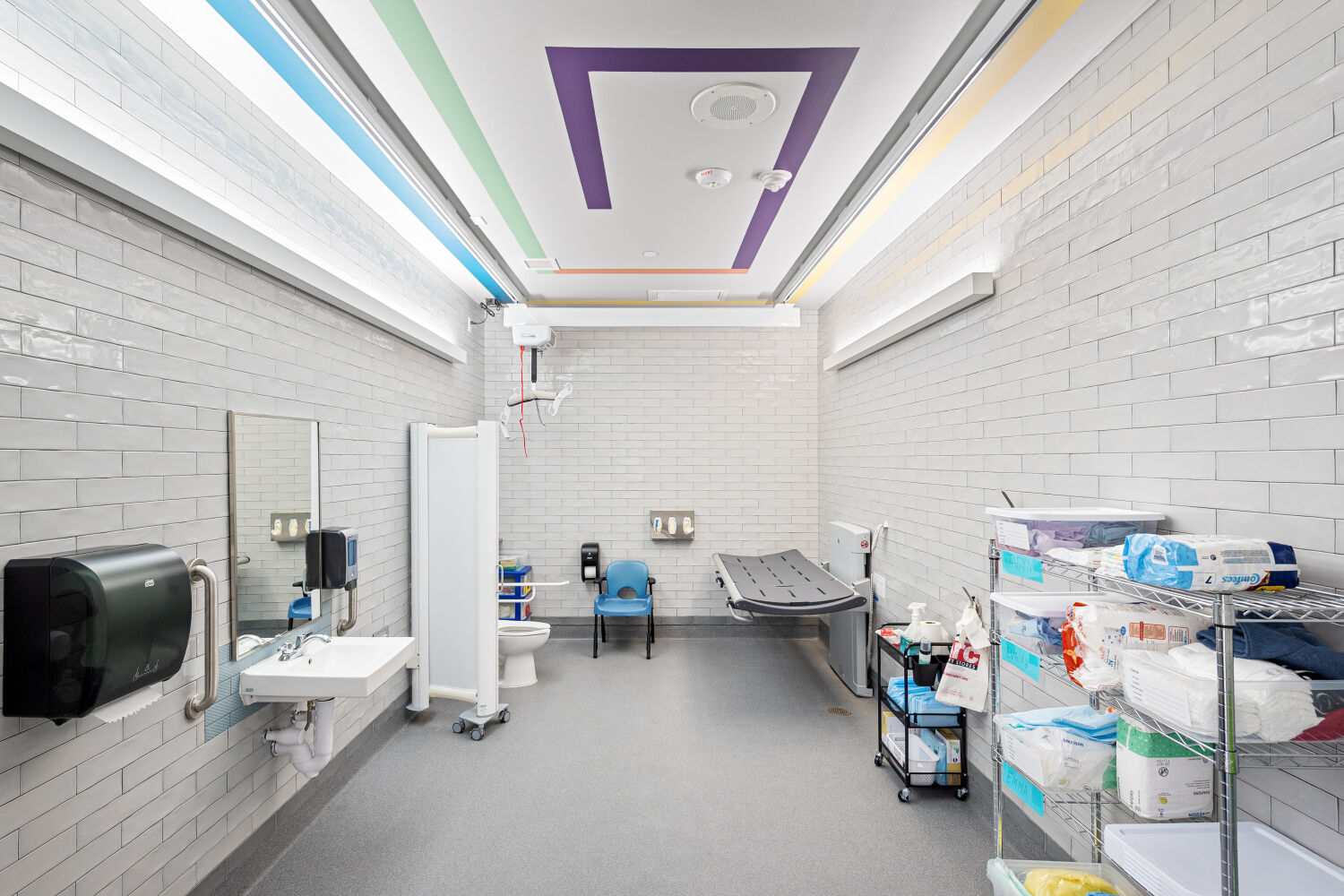 Hygiene room with a height‑adjustable care table, wall‑mounted sink, toilet with privacy screen, open storage shelving for supplies, and resilient flooring.