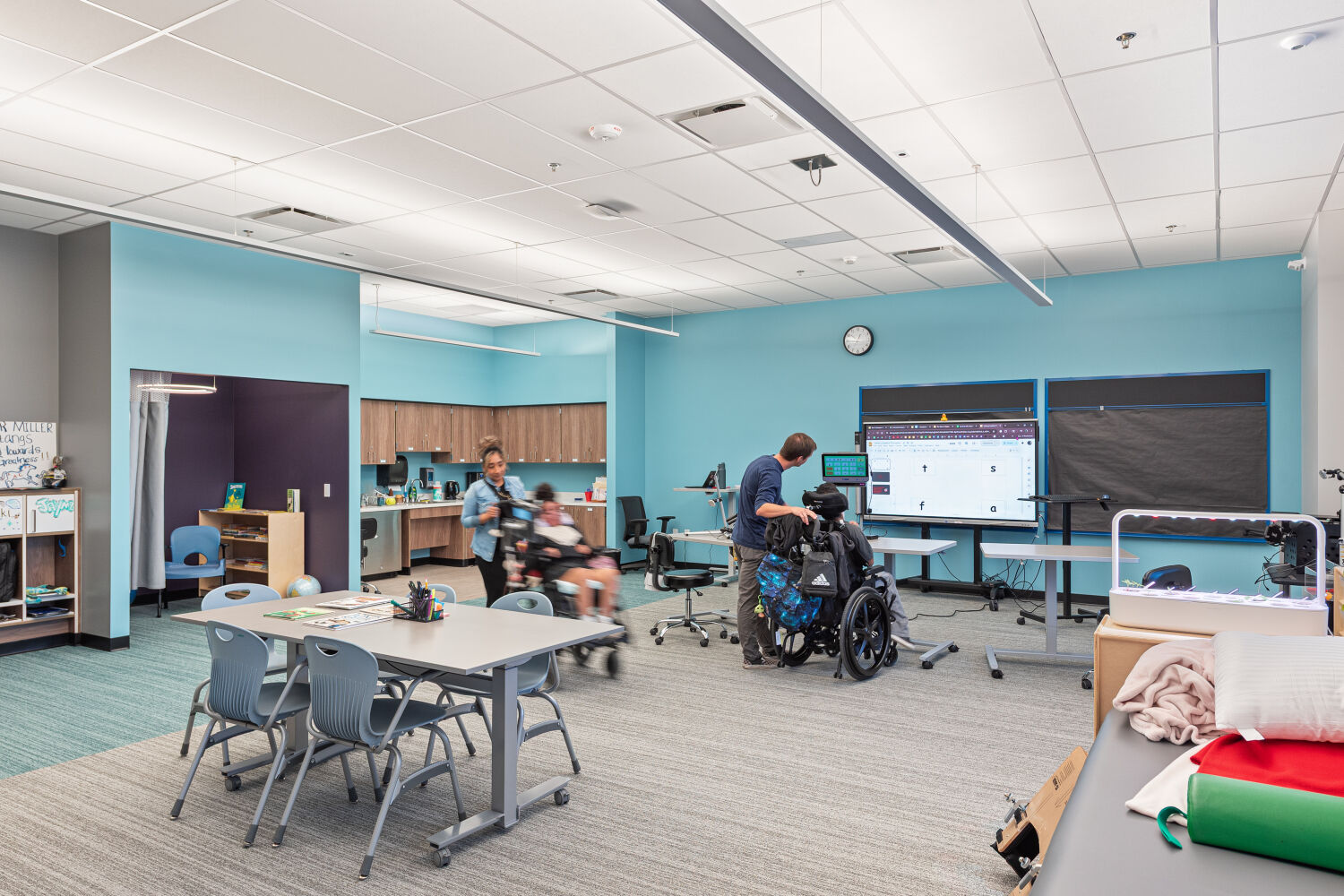 Specialized classroom with adjustable tables, mobile seating, assistive technology stations, wall‑mounted display screens, perimeter storage cabinets, and open instructional space.