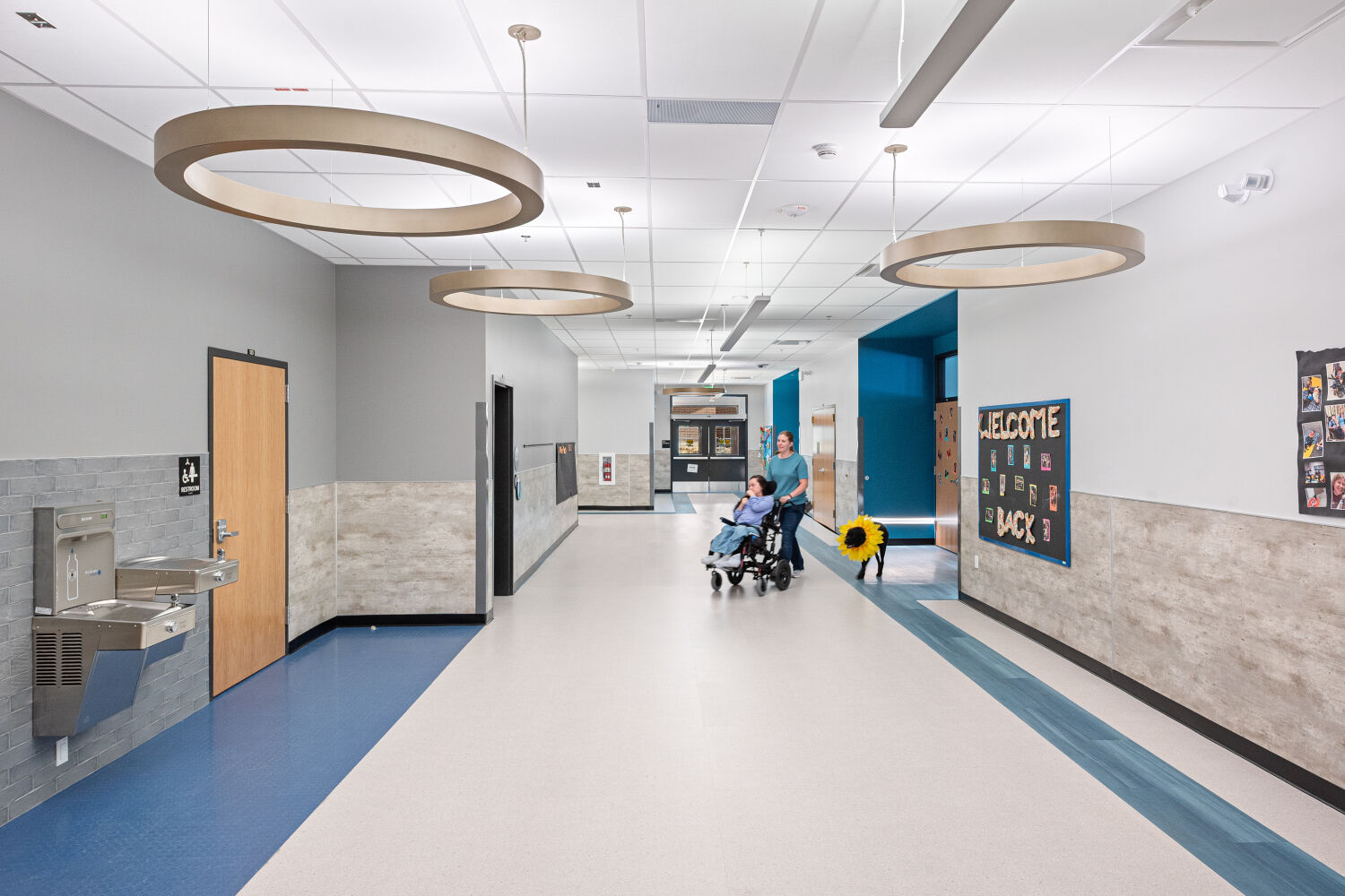 Learning corridor with wide circulation space, resilient flooring with color accents, circular suspended light fixtures, wall‑mounted drinking fountain, classroom doors, and student display boards.