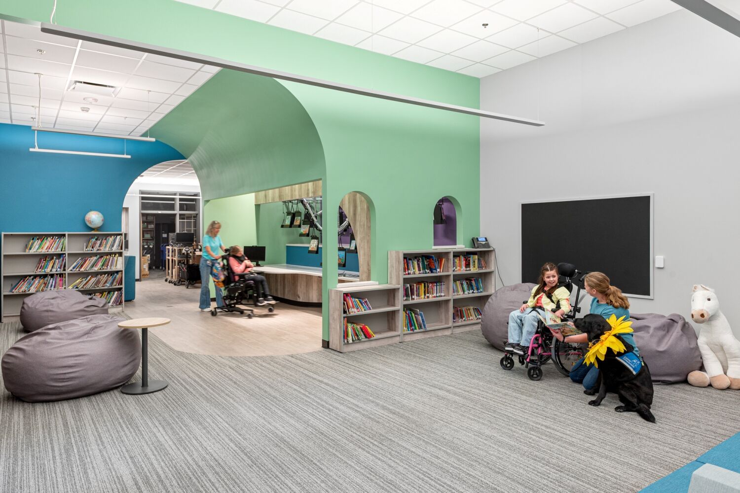 Learning commons with low library shelving, arched wall openings, soft seating, bean bags, resilient carpet flooring, and integrated ceiling lighting.