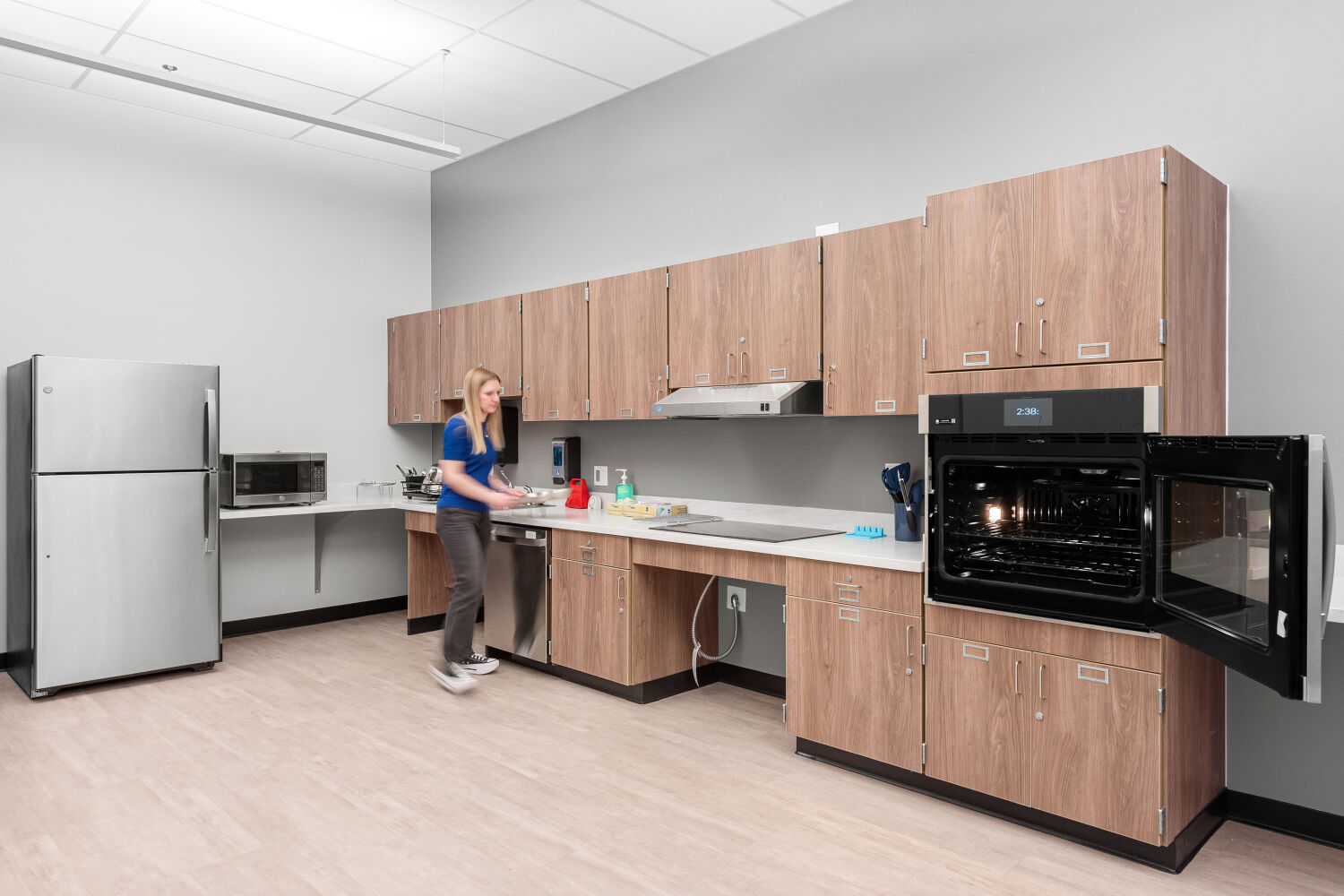 Life skills kitchen with perimeter cabinetry, refrigerator, microwave, wall oven, sink, continuous countertop workspace, and resilient flooring.