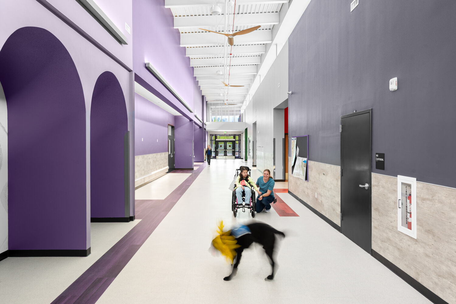 Main school corridor with arched wall openings, purple accent walls, resilient flooring with color inlays, high ceiling with exposed structure, and large ceiling fans.
