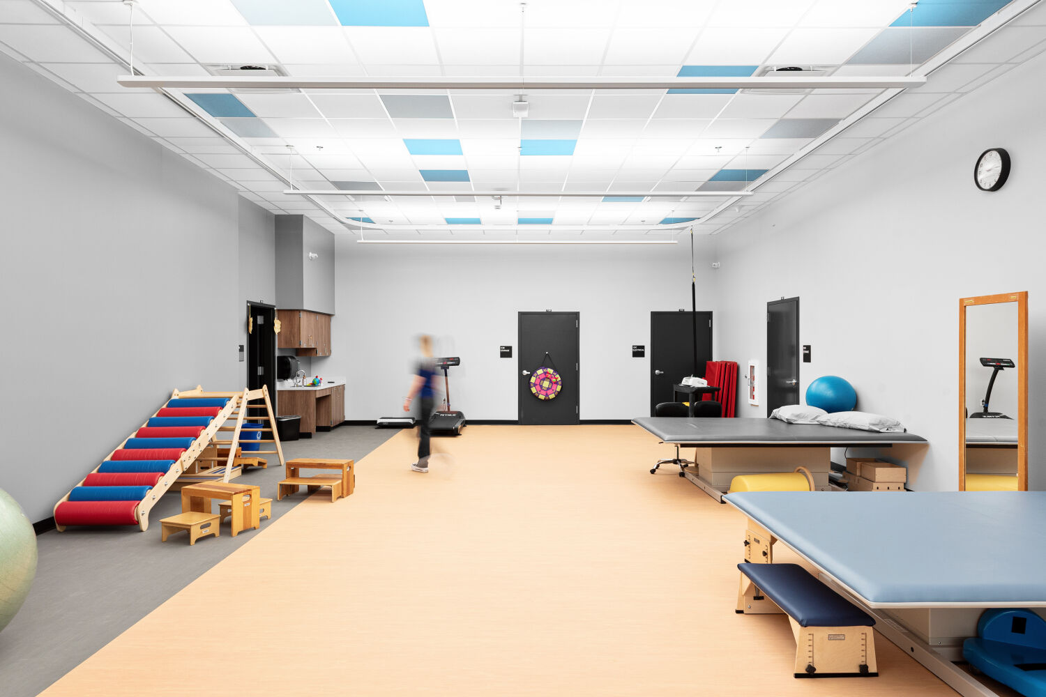 Physical therapy room with padded floor area, therapy tables, balance and climbing equipment, storage cabinetry, ceiling‑mounted tracks, and open space for movement.