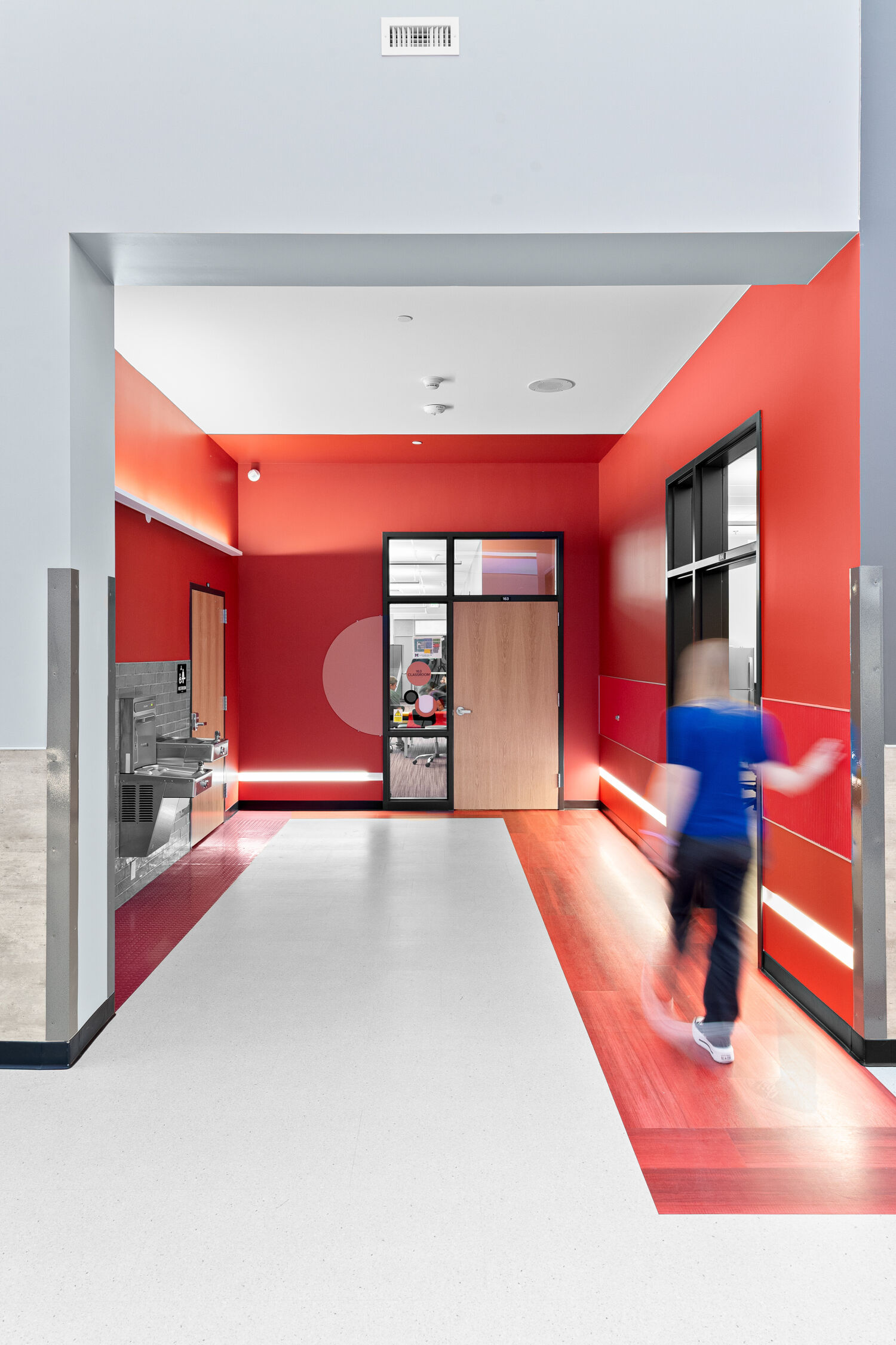 Learning corridor with red accent walls, resilient flooring with color inlays, a wall‑mounted drinking fountain, classroom glazing, and integrated linear lighting.