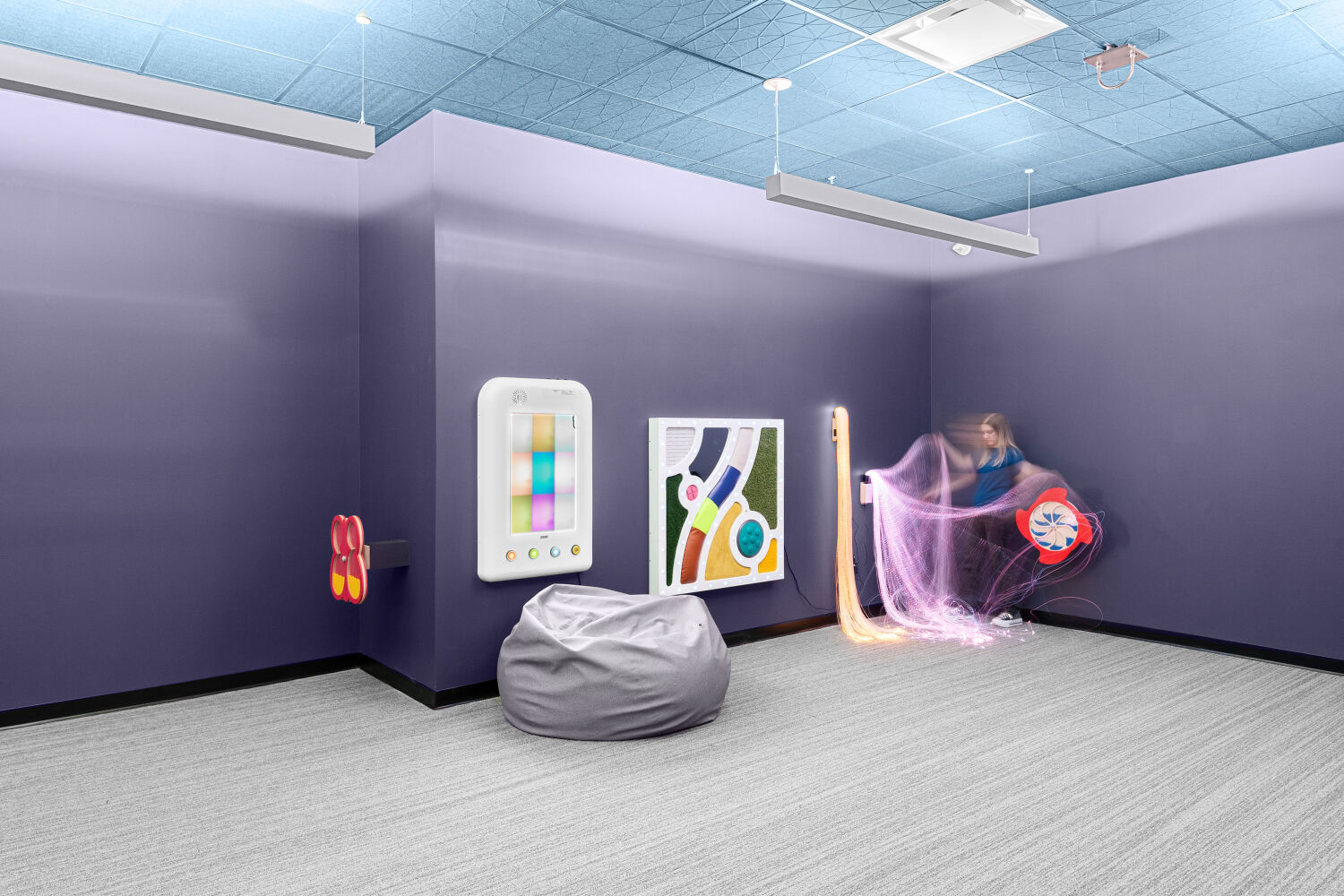 Sensory room with interactive wall panels, soft bean bag seating, resilient flooring, suspended acoustic ceiling panels, and adjustable linear lighting.