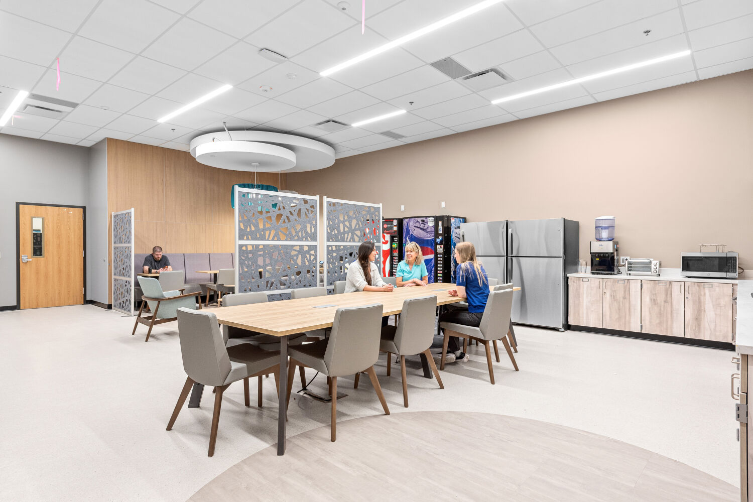 Teachers kitchen with a large communal table, upholstered seating, decorative privacy screens, kitchenette with appliances, pendant lighting, and adjacent quiet seating areas.