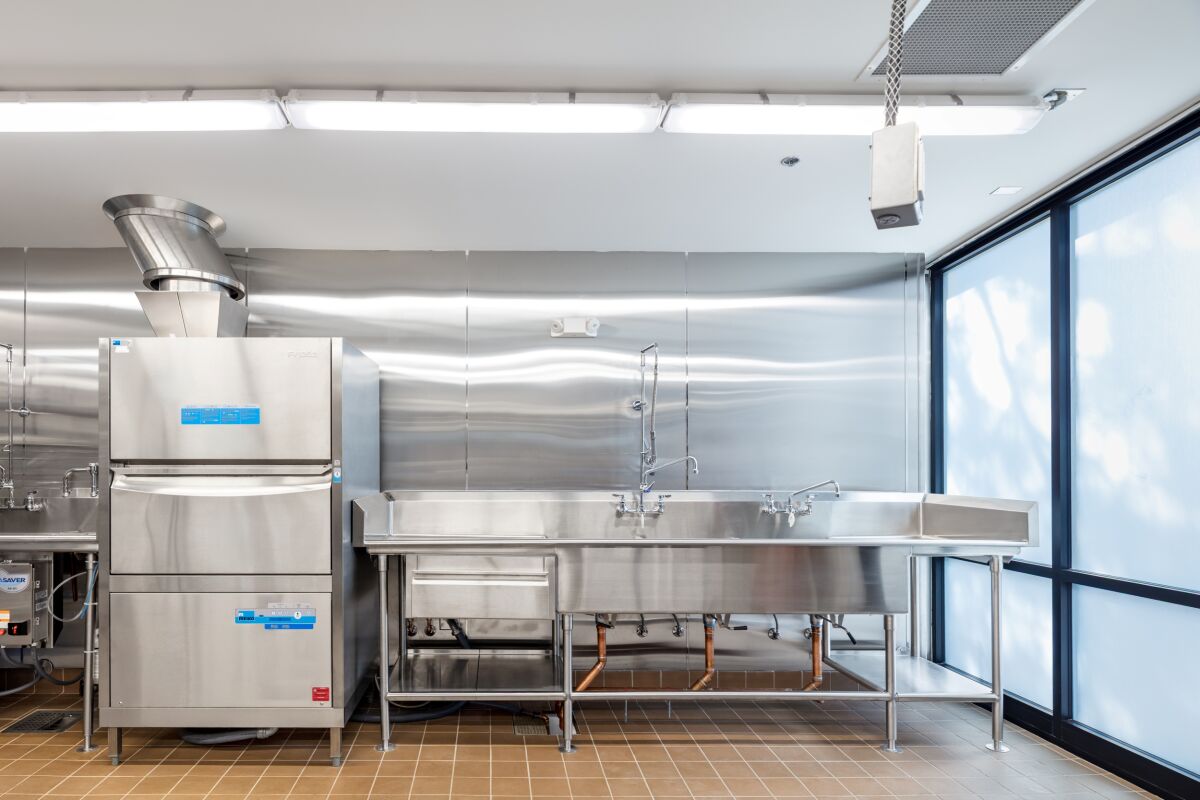 Commercial dishwashing area with a stainless‑steel dishwasher, triple‑compartment sink, overhead sprayer, metal wall paneling, tile flooring, and large frosted windows providing diffused natural light.