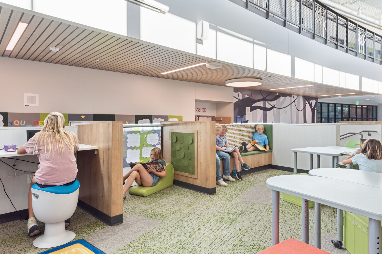 Media center with individual study pods featuring wood partitions and green cushioned seating, curved mezzanine walkway above, slatted ceiling panels, patterned carpet tile flooring, and surrounding tables and work areas.