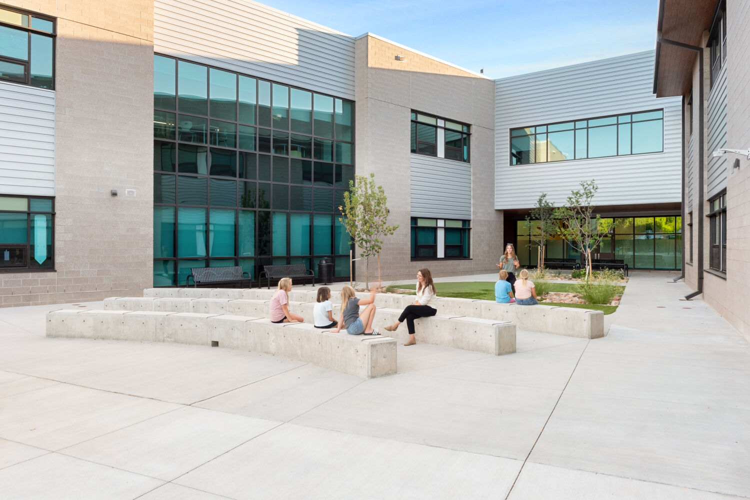 Outdoor learning courtyard enclosed by two‑story school buildings with large windows, featuring curved concrete seating walls arranged in a semicircle around a small planted area with young trees, surrounded by paved walkways.