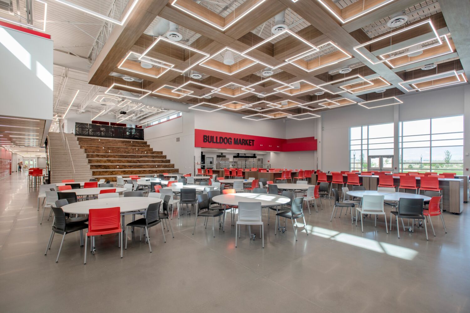 Cafeteria and commons area with round tables and chairs, a tiered learning stair, wood‑accent ceiling with geometric lighting, and a food‑service area labeled ‘Bulldog Market.’