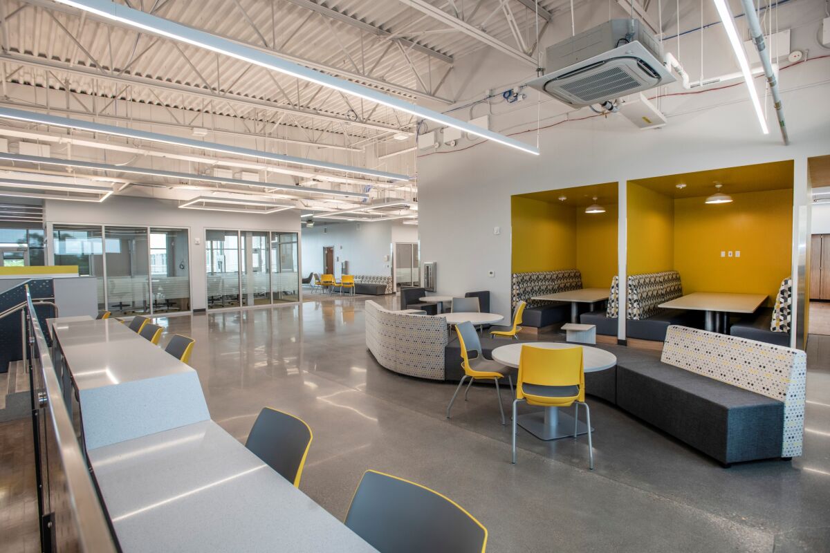 Collaboration area with lounge seating, round tables, booth-style study nooks, long worktables with yellow chairs, polished concrete flooring, and exposed ceiling structure with linear lighting.
