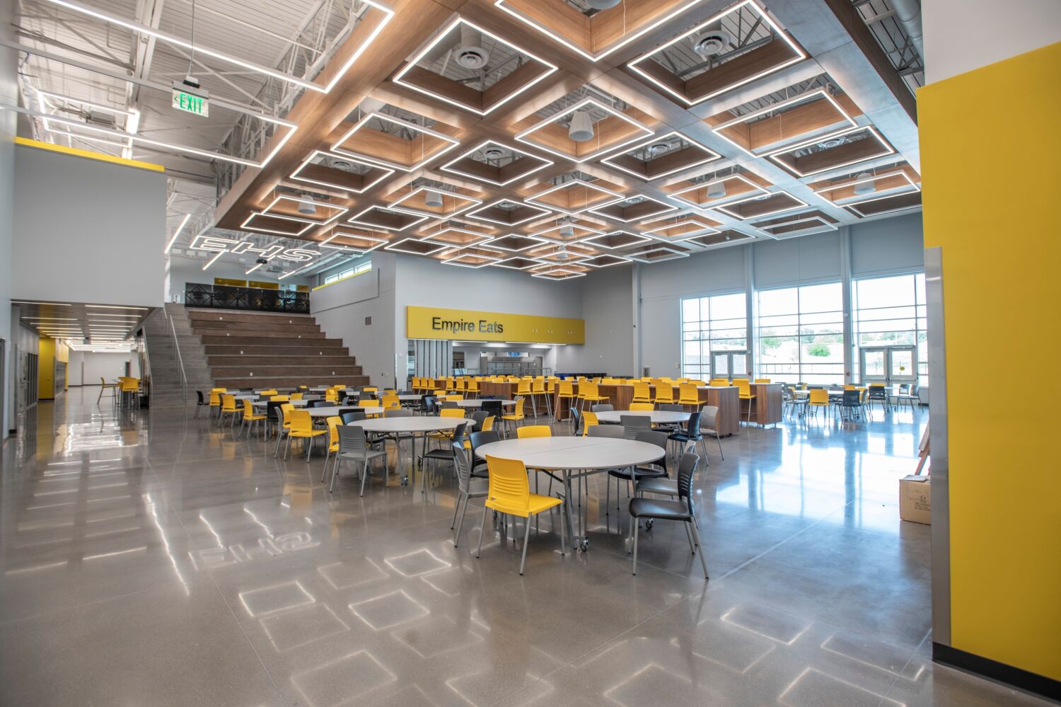 Cafeteria and commons with round tables and yellow chairs, a tiered learning stair, large windows, polished concrete flooring, and a wood‑accent ceiling with geometric LED lighting.