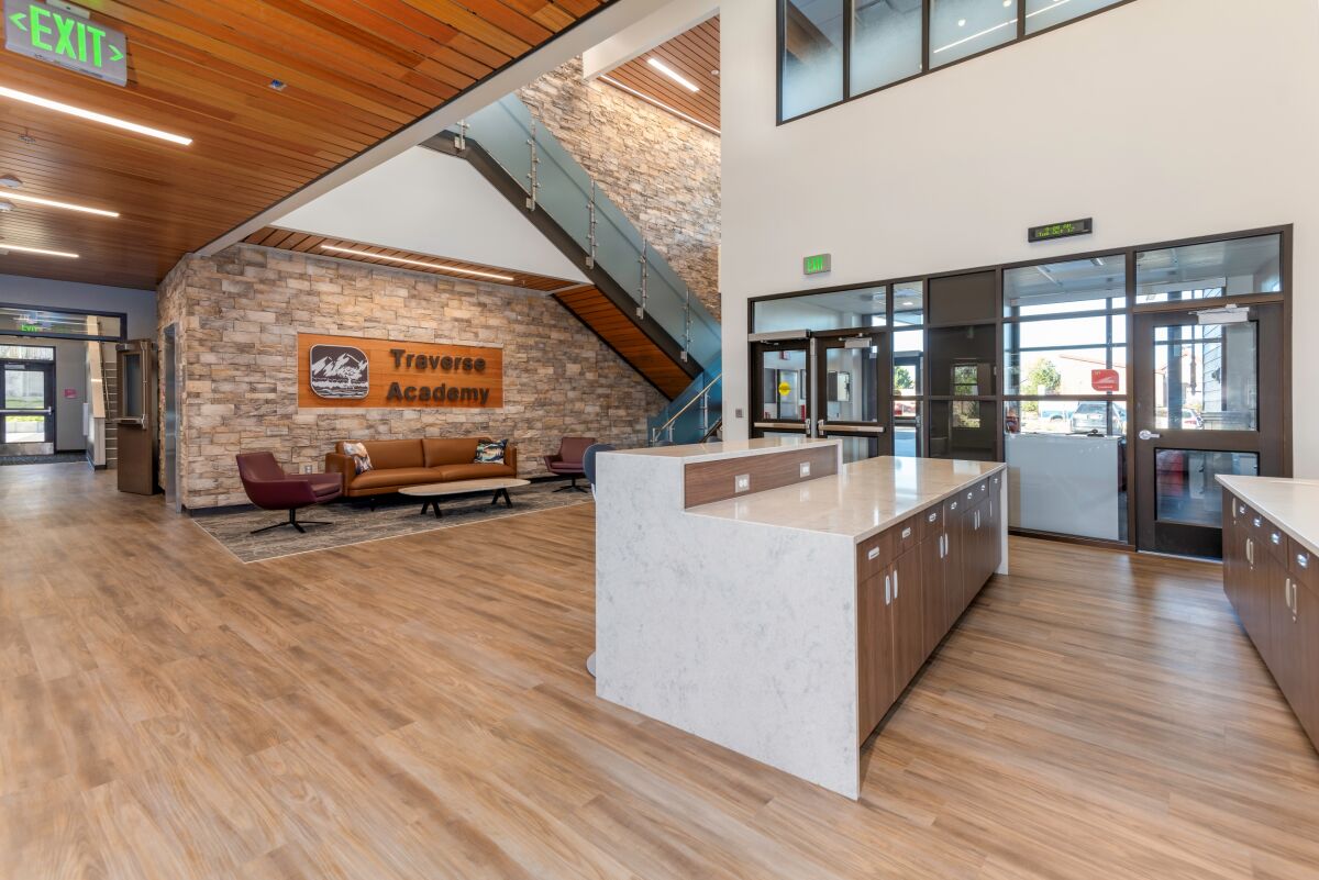 Café‑style entrance with a central counter, wood‑slat ceiling, stone feature wall with the Traverse Academy sign, lounge seating, and a glass‑railed staircase.