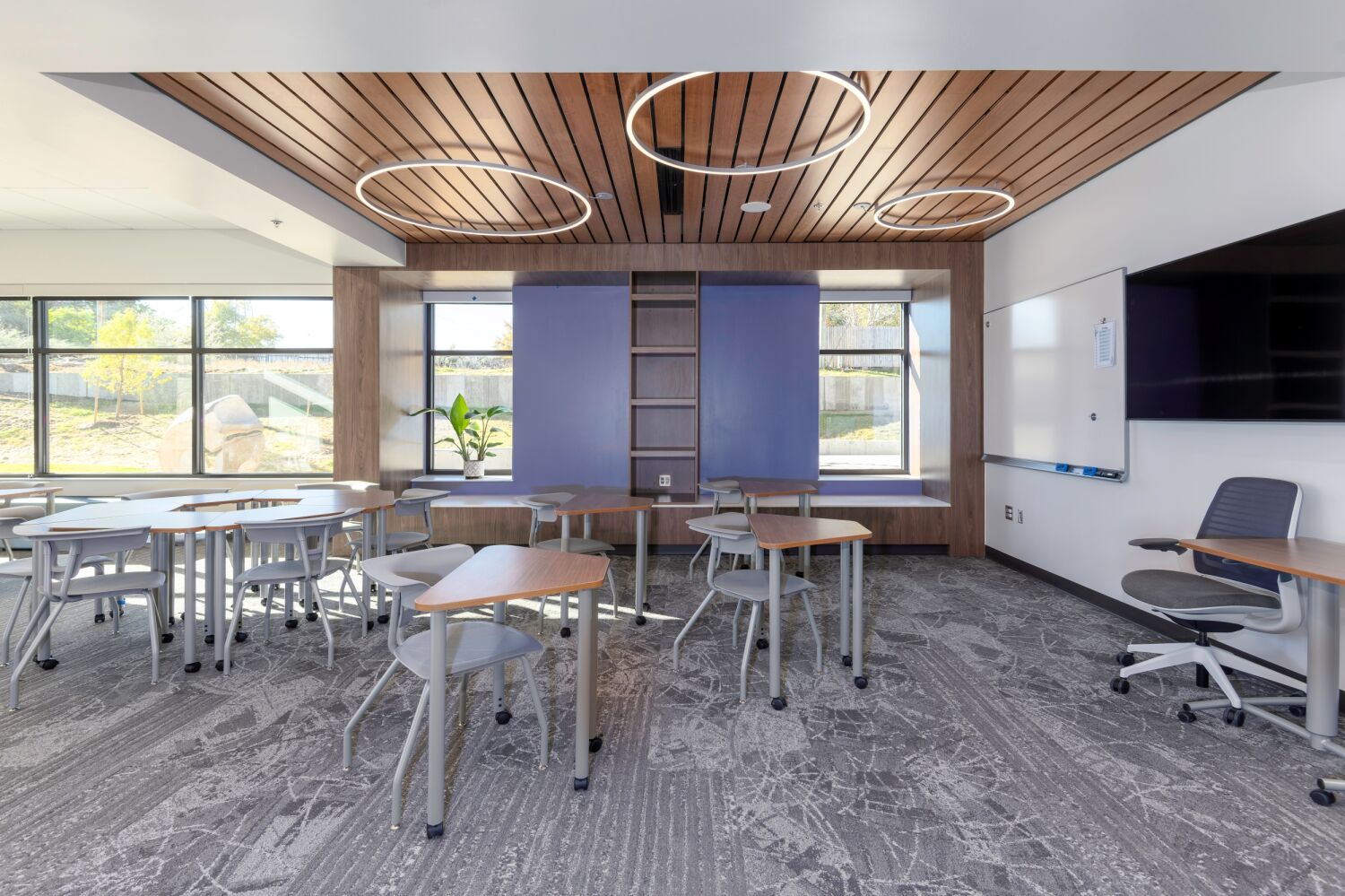 Classroom with modular tables and chairs, wood‑slat ceiling feature with circular lights, shelving niche with blue panels, and large windows.