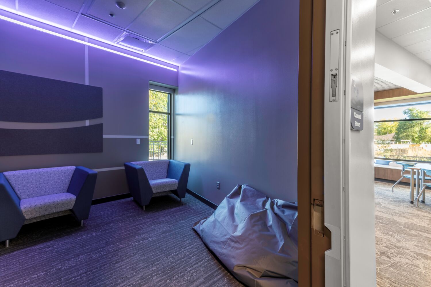 Sensory room with soft seating, acoustic wall panels, dim purple accent lighting, a beanbag pad on the floor, and a window providing natural light.