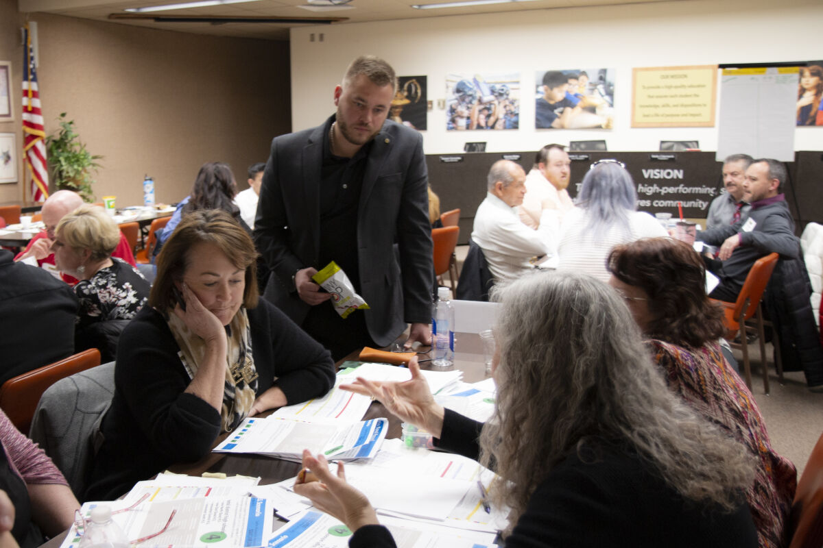 Community workshop with participants seated at tables reviewing planning documents, with facilitators moving between groups in a meeting room displaying posters and presentation boards.