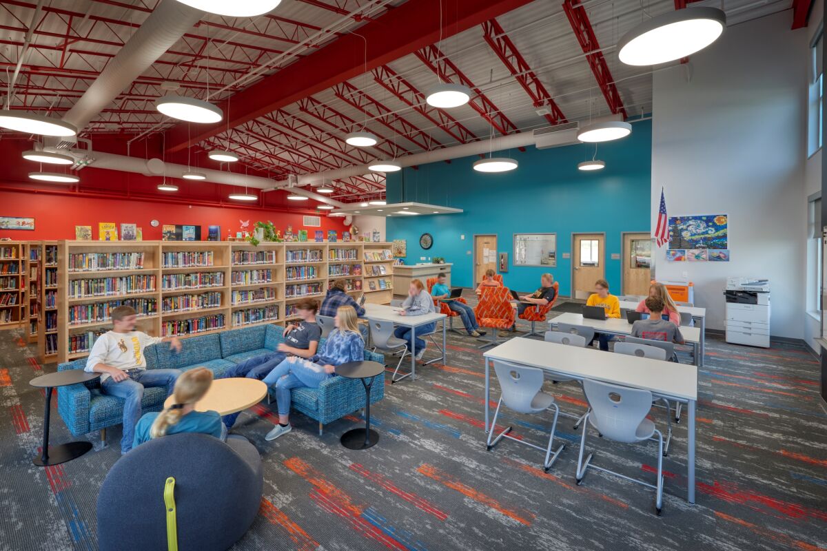 Library with bookshelves, lounge seating, study tables, colorful carpet patterns, red ceiling structure, and large circular light fixtures.