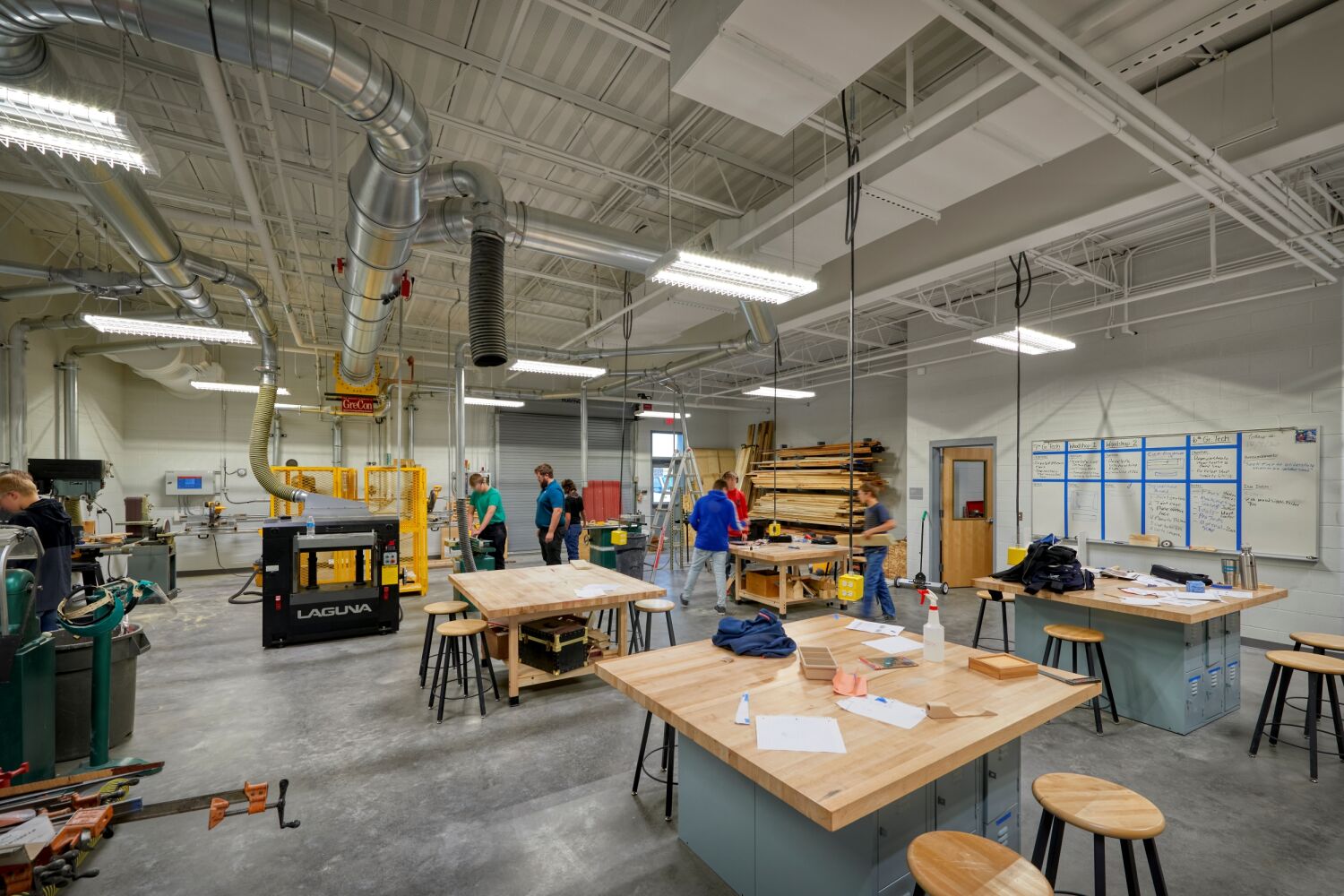 Wood shop with workbenches, stools, woodworking equipment, dust‑collection ducts, lumber storage racks, and whiteboard walls with project notes.