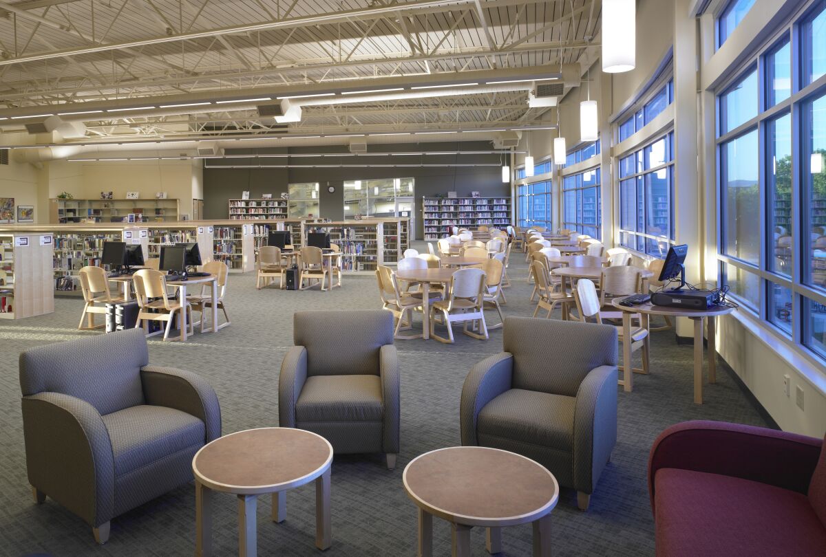 Library with rows of bookshelves, computer stations, wooden tables and chairs, lounge seating at the foreground, large windows along one wall, and an exposed ceiling with suspended lighting.