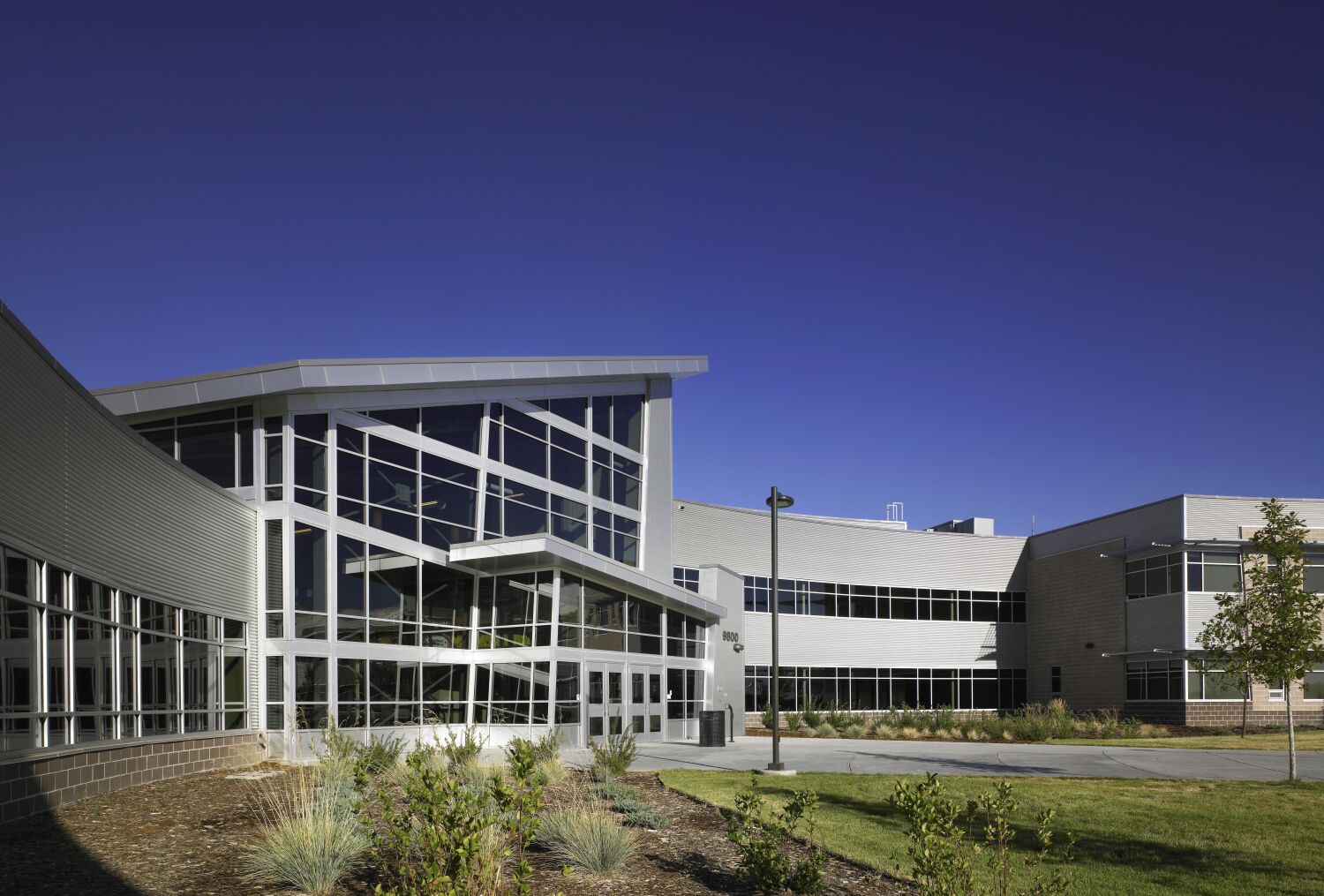 Exterior entrance of Bear Creek High School with a large glass façade, angled roofline, landscaped plantings, and a walkway leading to the main doors.