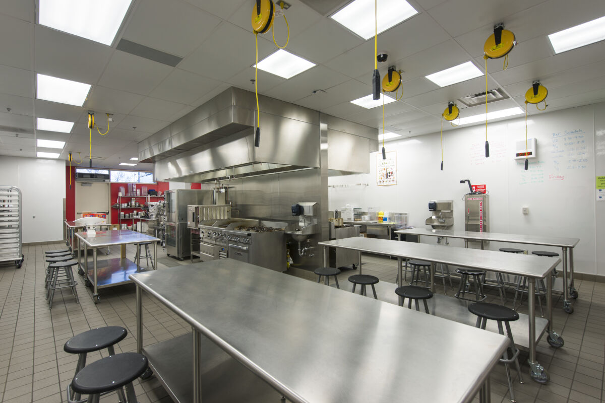 Culinary arts kitchen with stainless-steel worktables, stools, commercial cooking equipment, overhead ventilation hood, tile flooring, and ceiling-mounted power reels.