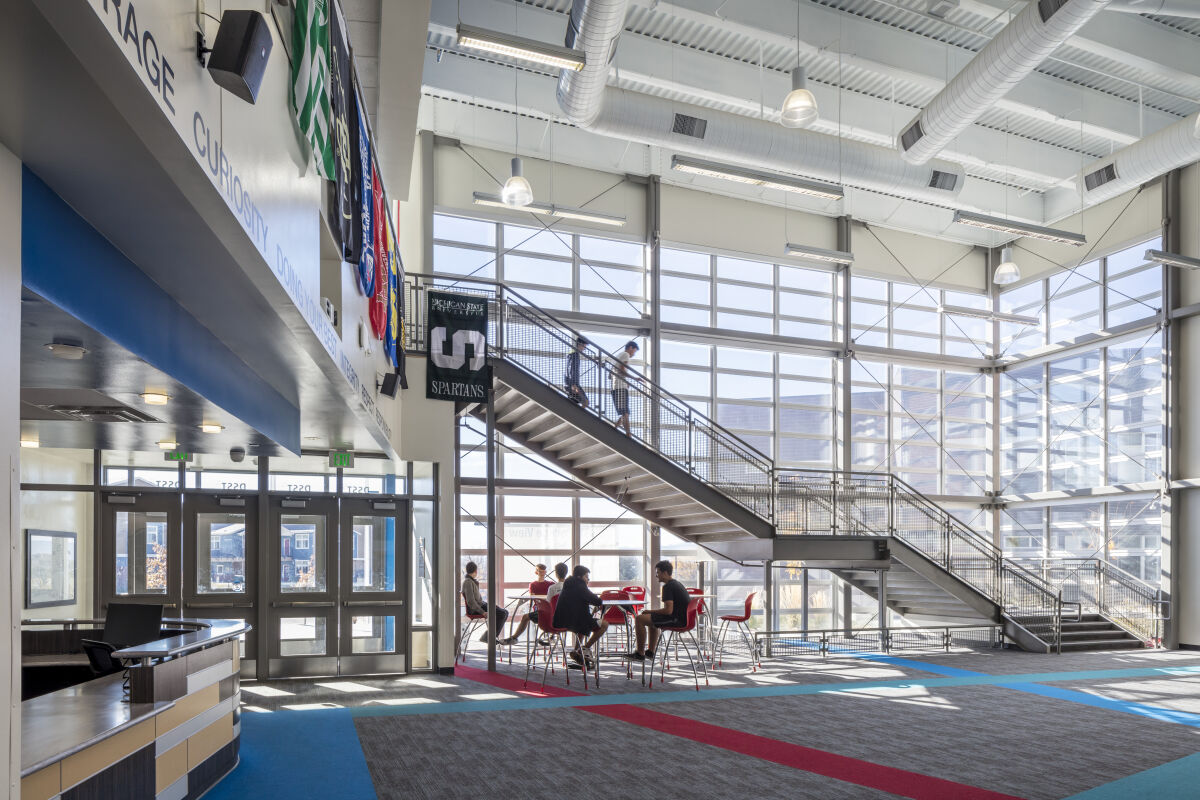 Lobby with a large glass curtain wall, open staircase, high ceilings with exposed ductwork, pendant lighting, entry doors, and seating at round tables on patterned carpet.