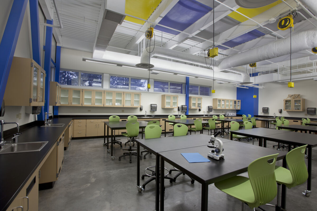 Science lab with black‑top worktables, green chairs, storage cabinets with glass doors, overhead power reels, exposed ceiling structure with ductwork, and windows along one wall.