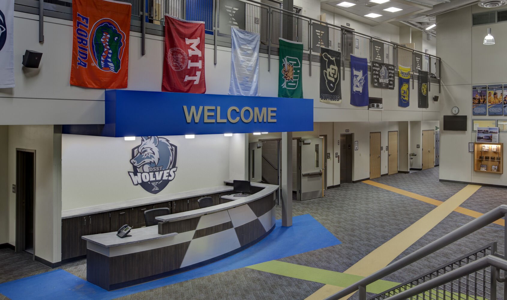Lobby with a curved reception desk under a blue ‘WELCOME’ sign, school banners hanging from the upper level, carpet tile flooring with colored accents, and classroom doors along the corridor.