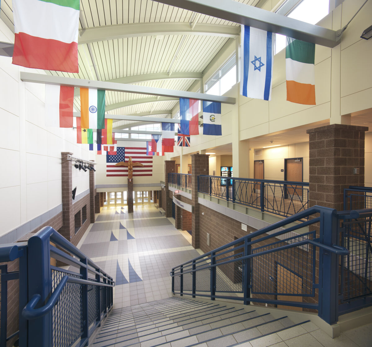 Two‑story commons with tiled flooring, central staircase, international flags hanging from the ceiling, brick columns, balcony railing on the upper level, and natural light from clerestory windows.