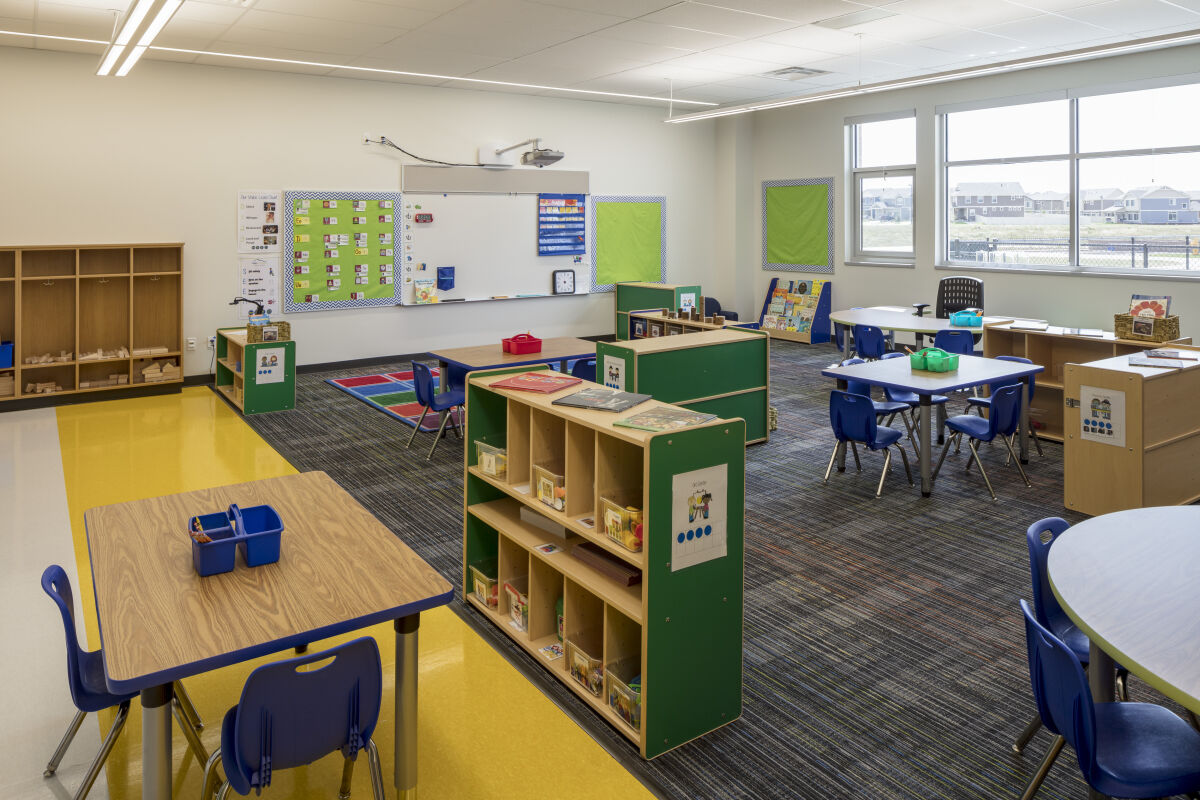 Early‑learning classroom with activity tables and chairs, open shelving units, a rug area near the whiteboard, colorful bulletin boards, and large windows providing natural light.